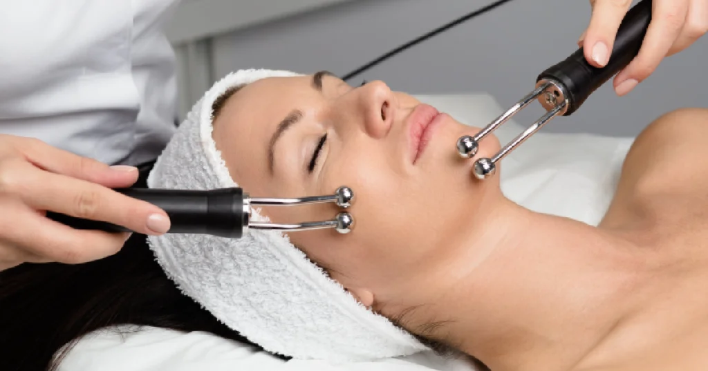 Woman receiving a professional microcurrent therapy treatment, lying relaxed with eyes closed while a practitioner uses dual metal probes on her cheeks in Niceville, FL